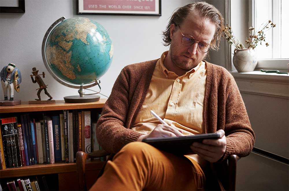 Bendik Kaltenborn bei der Arbeit an einem Tablet in seinem gemütlichen Arbeitszimmer, umgeben von Büchern und einem Globus. Fotografiert von Lars Petter Pettersen.