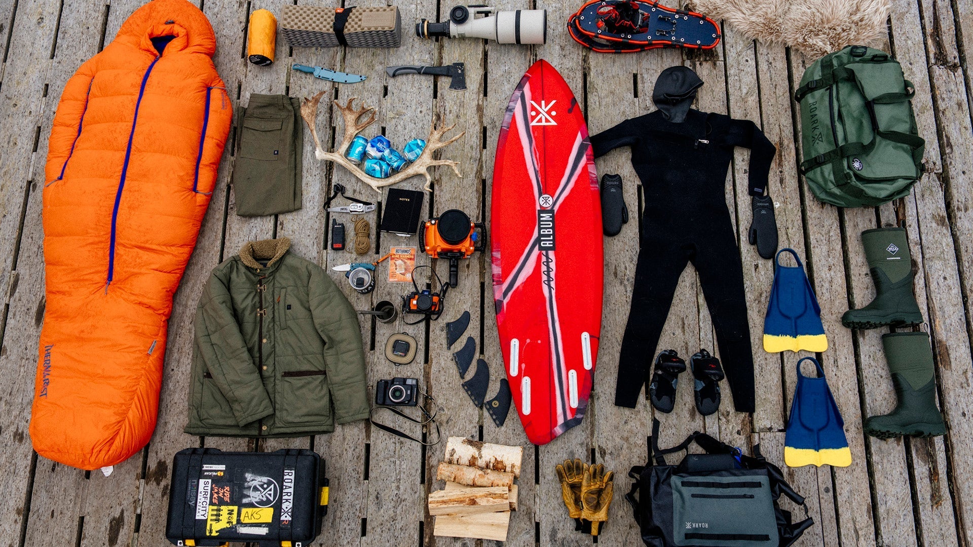 Eine flache Anordnung der wichtigsten Wintersurfausrüstung auf einem Holzsteg. Dazu gehören ein leuchtend orangefarbener Schlafsack, ein Neoprenanzug, ein Surfbrett, Winterkleidung und verschiedene Campingwerkzeuge.